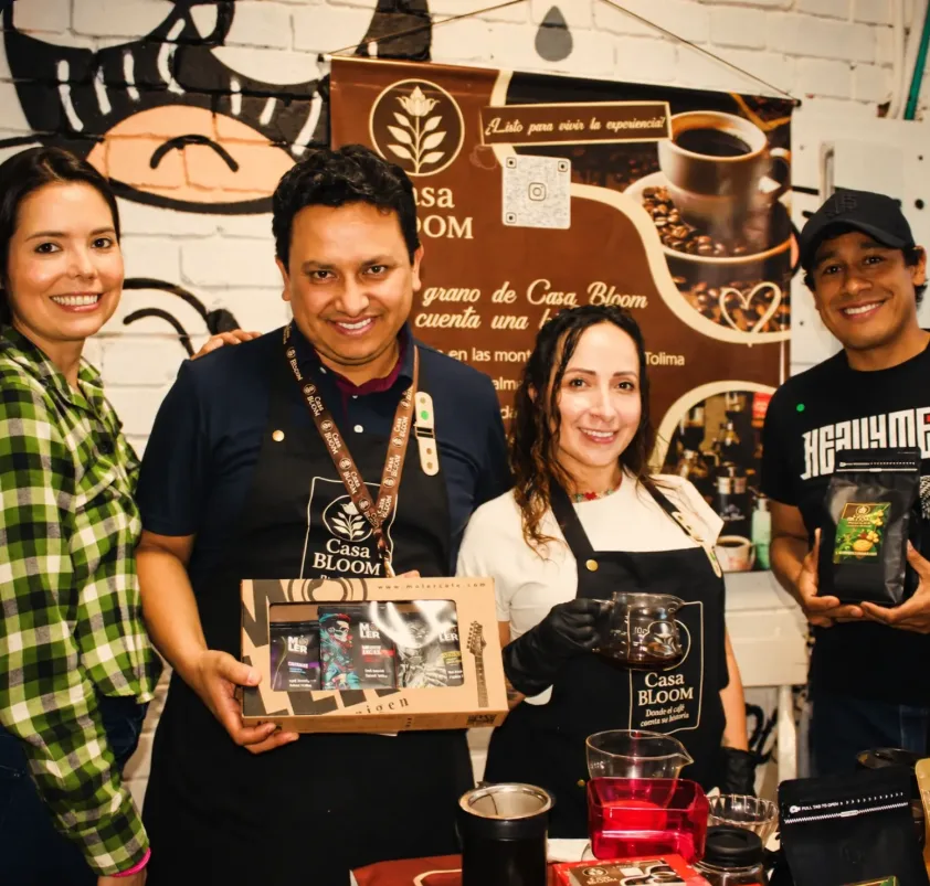 Equipo de Casa Bloom con delantales de la marca, sosteniendo café y una caja de regalo frente al pendón de Casa Bloom